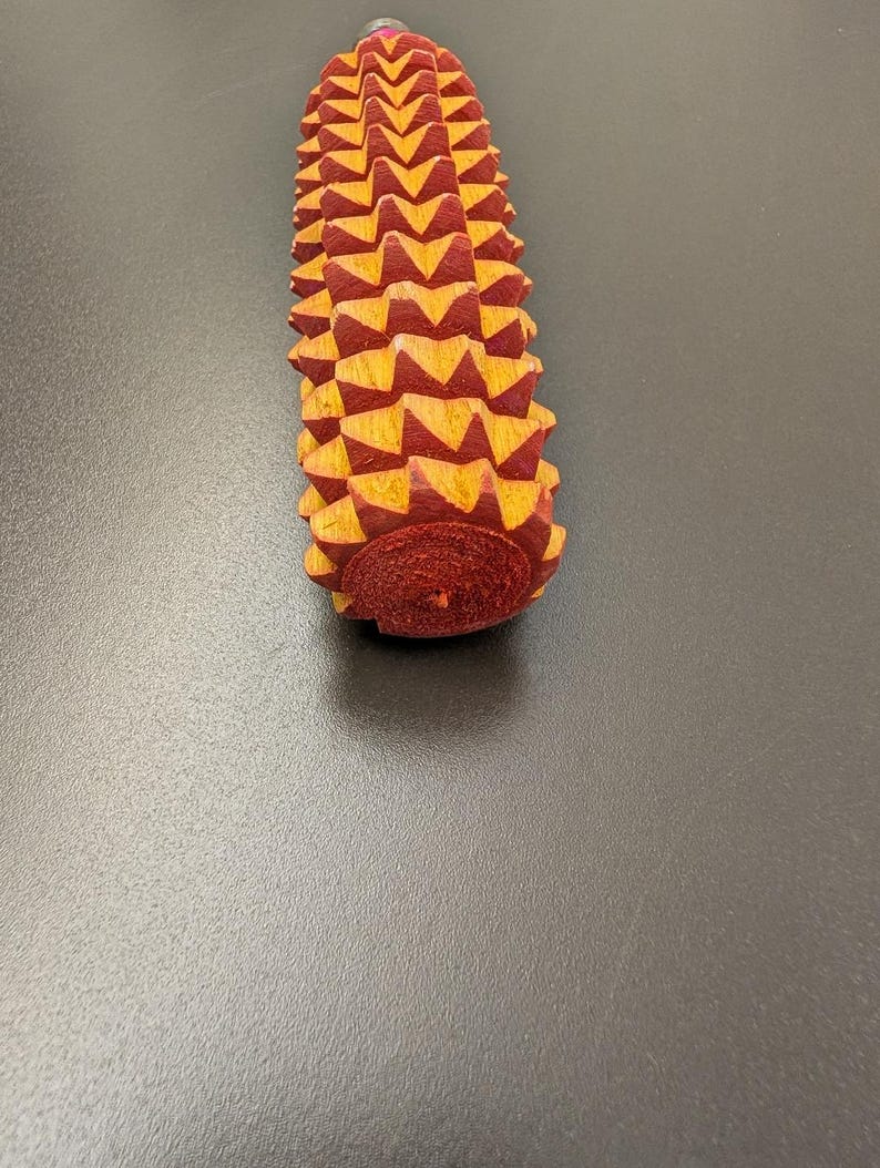 May include: A wooden massage tool with a textured surface. The tool is primarily red with yellow accents, and has a rounded base. The massage tool is designed to be held in the hand and used to massage muscles.