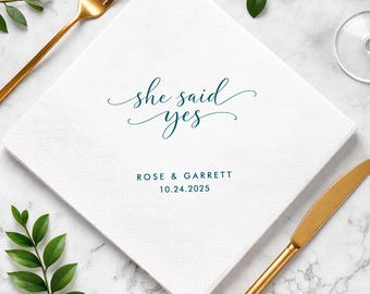 May include: A square white paper napkin featuring the dark teal text "she said yes" in a cursive script, followed by the names "ROSE & GARRETT" and the date "10.24.2025" printed in a simple, clean, dark teal sans-serif font.