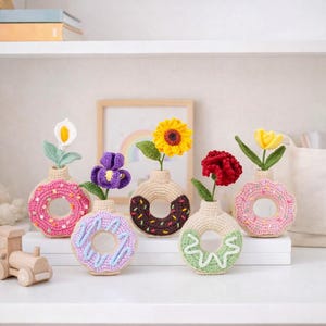 May include: Six crochet donut-shaped vases, each with a different crochet flower. The donuts are in various colours, including pink, purple, brown, and green, with white or pastel-coloured frosting details. The flowers include a sunflower, iris, and calla lily.