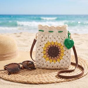 May include: A cream-colored crochet bucket bag featuring a central sunflower motif with brown and yellow petals, a green drawstring cord, and a small green leaf charm. The bag has a brown leather-like shoulder strap and gold-tone metal hardware.