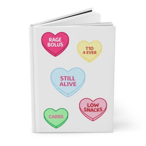 Puede incluir: Un cuaderno blanco con gráficos en forma de corazón de colores. Los corazones son rosas, amarillos, azules y verdes, cada uno con una frase diferente: "RAGE BOLUS", "T1D 4 EVER", "STILL ALIVE", "LOW SNACKS" y "CARBS".
