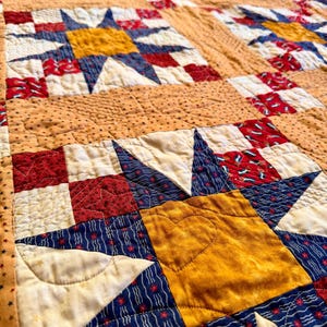 May include: Close-up of a handmade quilt with a star pattern. The quilt features a central yellow star with blue and red accents, set against a cream and tan background. The quilt is made of fabric and has a quilted texture.