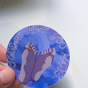 Könnte beinhalten: Ein runder Aufkleber mit blau-violettem Himmelshintergrund, der zwei violette Füße zeigt, einer davon mit einem goldenen Ring an einem Zeh, sowie den Text "TELL ME WHAT YOU WANT" in weißer Schreibschrift am Rand des Aufkleberdesigns.