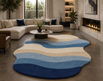 May include: An area rug with an irregular, organic shape featuring horizontal wavy bands of color. The design consists of alternating stripes in shades of deep navy blue, medium blue, light blue, and a soft cream beige, creating a fluid pattern.