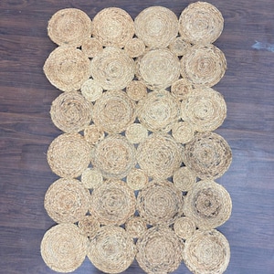 Peut inclure: Un tapis rectangulaire en jute avec un motif de cercles tissés de différentes tailles. Le tapis est de couleur beige naturel et repose sur un sol en bois foncé. Les cercles sont tissés serrés, créant une surface texturée.