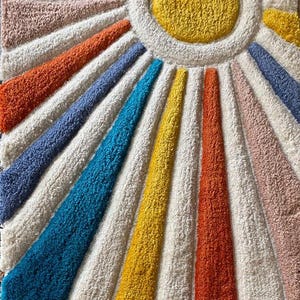 Könnte beinhalten: Ein farbenfroher, strukturierter Teppich mit einem Sonnenmuster. Der Teppich hat einen zentralen gelben Kreis, der von einem weißen Ring umgeben ist, mit Strahlen in verschiedenen Farben, darunter Orange, Blau und Rosa. Die Größe des Teppichs ist nicht erkennbar.