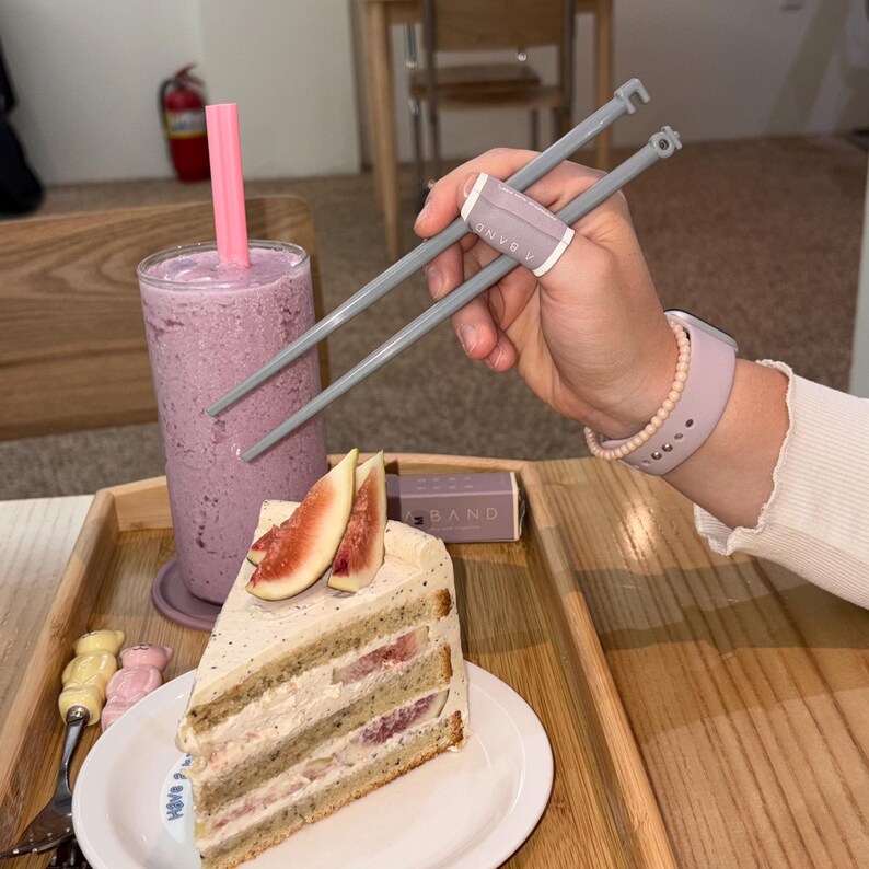 Puede incluir: Una rebanada de pastel en capas con rodajas de higos en un plato blanco, un batido morado en un vaso alto con una pajita rosa y palillos chinos grises con una banda rosa. Tambi&eacute;n se ve un reloj y una pulsera rosas.