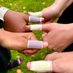Puede incluir: Primer plano de varias manos con vendas en los dedos. Las vendas son rectangulares, con una base de color lila claro o amarillo y ribete rosa. El texto "A BAND" es visible en una venda. El fondo es hierba verde borrosa.