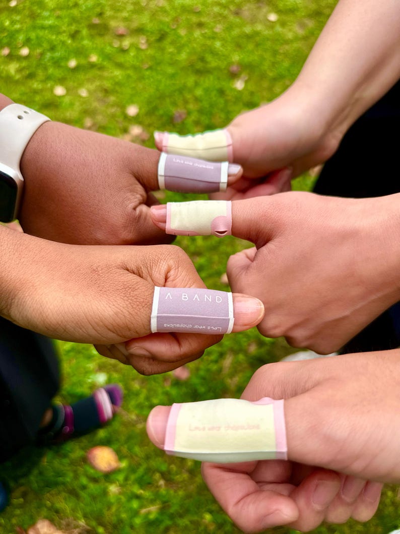 Puede incluir: Primer plano de varias manos con vendas en los dedos. Las vendas son rectangulares, con una base de color lila claro o amarillo y ribete rosa. El texto "A BAND" es visible en una venda. El fondo es hierba verde borrosa.