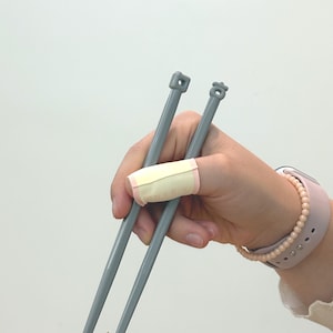 Puede incluir: Dos agujas de tejer grises se sostienen en una mano con una venda en el dedo &iacute;ndice. Las agujas tienen puntas cuadradas y en forma de flor. Un reloj rosa con una pulsera de perlas est&aacute; en la mu&ntilde;eca.