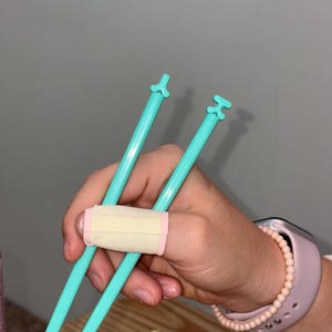 Puede incluir: Un par de palillos de entrenamiento azul claro con una banda de conexi&oacute;n. Los palillos tienen un dise&ntilde;o &uacute;nico en la parte superior. Una persona sostiene los palillos con una banda en el dedo.