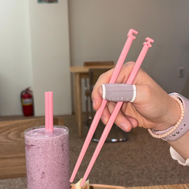 Puede incluir: Palillos chinos rosas con una banda lavanda se sostienen sobre un vaso de batido morado con una pajita rosa. Los palillos tienen una forma &uacute;nica en la parte superior. La banda tiene la palabra "BAND" impresa.