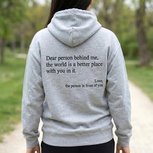 Könnte beinhalten: Ein grauer Kapuzenpullover mit dem Text "Dear person behind me, the world is a better place with you in it. Love, the person in front of you" auf dem Rücken. Der Kapuzenpullover hat eine Kapuze und lange Ärmel.