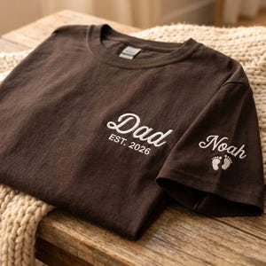 May include: A dark brown t-shirt featuring white embroidery on the chest that reads Dad EST. 2026. The sleeve is embroidered with the name Noah in white script lettering accompanied by a small white graphic of two baby footprints.