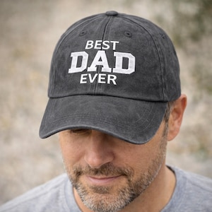 May include: A charcoal gray baseball cap with the words "BEST DAD EVER" in white block letters. The cap has a curved brim and a classic six-panel design. The hat is made of a soft, worn material.