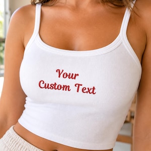 Könnte beinhalten: Ein weißes, geripptes Tanktop mit dünnen Trägern und einem Rundhalsausschnitt. In der Mitte der Brust befindet sich der in Rot gestickte Text "Your Custom Text" in Schreibschrift, was einen klaren Blick auf die Textur des Kleidungsstücks ermöglicht.