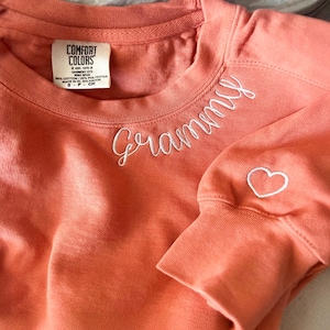 May include: A coral-coloured long-sleeve sweatshirt featuring white embroidered cursive text that reads Grammy on the chest. A small white embroidered heart is located on the sleeve. The interior neck label reads Comfort Colors, 80% cotton, 20% polyester.