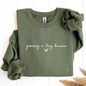 Könnte beinhalten: Ein olivgrünes Sweatshirt mit dem weißen Schriftzug "growing a tiny human" und einem kleinen Herz. Die langen Ärmel sind gefaltet. Das Gildan-Markenetikett ist sichtbar.