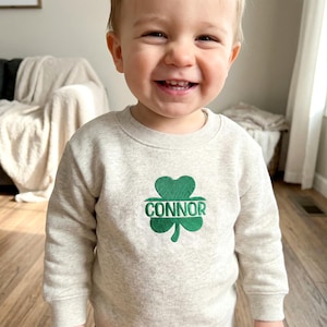 May include: A light gray sweatshirt with a green embroidered shamrock and the name "Connor" in green lettering. The sweatshirt is a crew neck style and appears to be made of a soft material. The design is centered on the front of the shirt.
