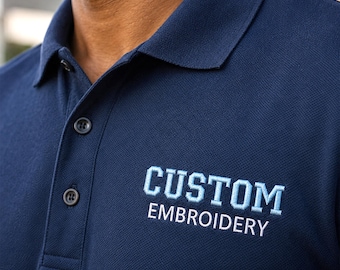 Puede incluir: Un polo azul marino de tejido piqué texturizado, con cuello, tapeta con tres botones oscuros y las palabras CUSTOM EMBROIDERY bordadas en el pecho en letras de bloque de color azul claro y blanco.