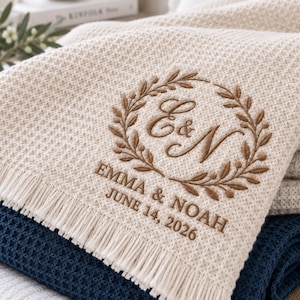 May include: A cream-coloured waffle-knit throw blanket with fringe edges, featuring brown embroidery of a laurel wreath surrounding the initials E & N, with the text EMMA & NOAH JUNE 14, 2026, layered over a dark navy blue waffle-knit blanket.