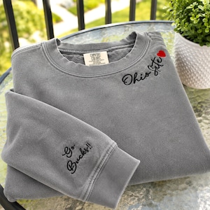 May include: A grey crewneck sweatshirt featuring the embroidered black text "Ohio Life" with a small red heart on the upper chest. The sleeve displays the embroidered black text "Go Bucks!!". The interior neck label reads "Comfort Colors".