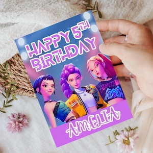 Puede incluir: Tarjeta de cumpleaños con el texto "HAPPY 5TH BIRTHDAY AMELIA" en morado. La tarjeta presenta tres personajes femeninos animados con cabello y ropa vibrantes sobre un fondo azul y rosa. La tarjeta es sostenida por una mano.