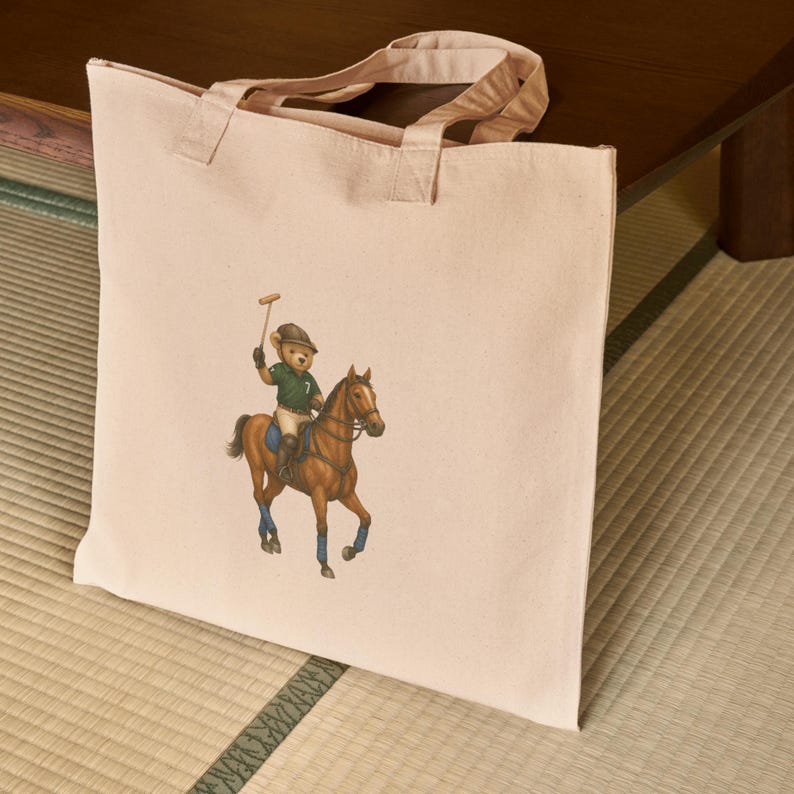 May include: A pale pink tote bag with a beige handle. The bag features a cartoon bear playing polo on a brown horse. The bear wears a green shirt, a brown helmet, and blue riding boots. The horse has blue leg wraps.