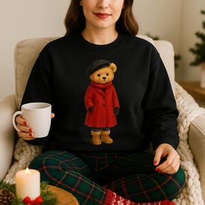 K&ouml;nnte beinhalten: Schwarzes Sweatshirt mit einer Teddyb&auml;r-Illustration. Der B&auml;r tr&auml;gt einen roten Mantel, eine schwarze Baskenm&uuml;tze und Stiefel. Die Person im Bild h&auml;lt eine wei&szlig;e Tasse und tr&auml;gt eine karierte Pyjamahose.