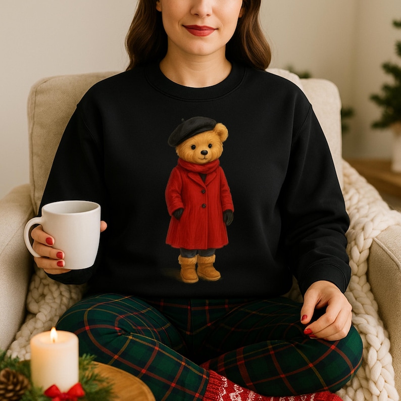 K&ouml;nnte beinhalten: Schwarzes Sweatshirt mit einer Teddyb&auml;r-Illustration. Der B&auml;r tr&auml;gt einen roten Mantel, eine schwarze Baskenm&uuml;tze und Stiefel. Die Person im Bild h&auml;lt eine wei&szlig;e Tasse und tr&auml;gt eine karierte Pyjamahose.