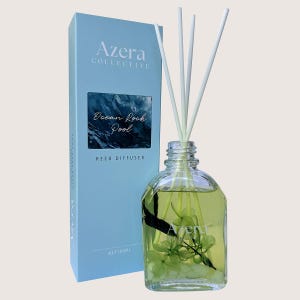 May include: A reed diffuser with a clear glass bottle containing a light green liquid and white reeds. The bottle is next to a light blue box with the text "Azera Collective" and "Ocean Rock Pool" printed on it. The bottle contains 100ml of liquid.