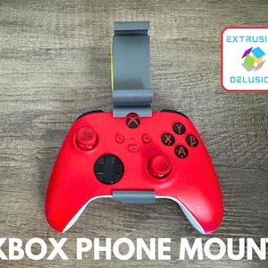 May include: A red Xbox controller with a gray phone mount attached. The controller has black buttons and white accents. The text "XBOX PHONE MOUNT" is at the bottom of the image. The Extrusion Delusion logo is in the top right corner.