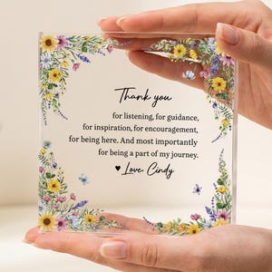 Può includere: Targa in acrilico trasparente con decorazioni floreali e la scritta "Thank you" seguita da un messaggio personalizzato. La targa è tenuta tra le mani di una persona. Il design floreale comprende girasoli gialli, fiori viola e blu.