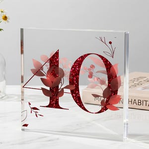 May include: A square clear acrylic table number sign featuring the number 10 in a deep red glitter finish. The design is accented with delicate red leaf and berry illustrations layered behind and around the bold, sparkling numerals.