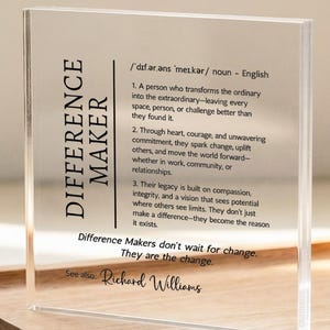Op de afbeelding: Een helder acrylblok met de woorden "DIFFERENCE MAKER" verticaal afgedrukt aan de linkerkant. Het blok bevat tekst die "Difference Maker" definieert en een citaat. Een kleine ingelijste foto en een laptop staan op de achtergrond.