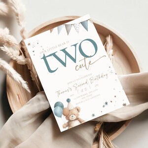 Puede incluir: Una invitación de cumpleaños con el texto "OUR LITTLE BEAR IS TWO cute". La invitación presenta una ilustración de oso de peluche, globos y una pancarta. La invitación está sobre un fondo blanco con una bandeja de madera.