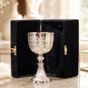 Può includere: Un calice in argento lucido con intricati motivi in rilievo sulla coppa e sulla base, posizionato davanti a una scatola di presentazione aperta foderata in velluto nero con chiusure in metallo dorato sui lati del contenitore rettangolare.