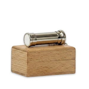 Puede incluir: Un caleidoscopio cilíndrico de metal plateado con una pieza circular blanca en el extremo, colocado horizontalmente sobre una caja rectangular de madera de color marrón claro con textura de veta visible y una costura horizontal.