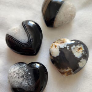 May include: Four heart-shaped stones with polished surfaces. The stones are primarily black and white, with variations in pattern and texture. Some have a banded appearance, while others have a speckled or crystalline look. The hearts are arranged on a white fabric.