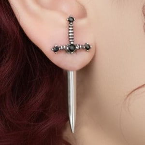 May include: A silver-colored sword earring with black gemstone accents. The earring features a detailed hilt with a vertical bar and a horizontal crossbar adorned with small, dark stones. The blade is long and slender, creating a unique and edgy accessory.