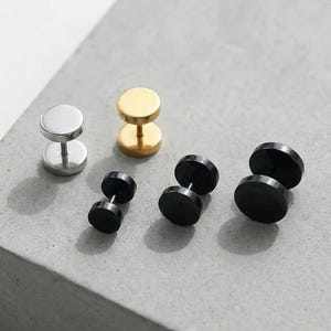May include: A collection of stud earrings in various colors. The earrings include silver, gold, and black options. The earrings have a flat, circular design and a post backing. The earrings are displayed on a gray surface.