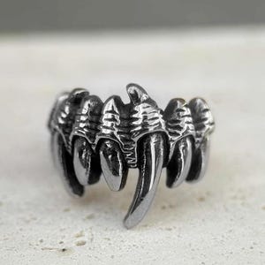 May include: A silver-toned ring with a claw design. The ring features multiple pointed teeth-like structures, creating a fierce and edgy aesthetic. The metal has a slightly darkened finish, enhancing the details of the claw design.