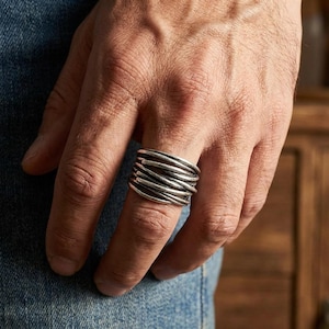 Puede incluir: Un anillo plateado con un diseño en capas. El anillo presenta múltiples bandas de metal envueltas entre sí, creando un aspecto texturizado. El anillo se lleva en un dedo, mostrando sus intrincados detalles y artesanía.