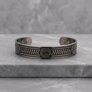 Könnte beinhalten: Ein silberfarbenes Armreif-Armband mit einem eingravierten Lebensbaum-Design. Das Armband weist ein sich wiederholendes Muster aus Blättern und kreisförmigen Emblemen auf. Das offene Design ermöglicht ein einfaches Tragen. Das Armband wird auf einer grauen Oberfläche präsentiert.