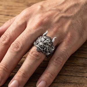 May include: A silver-colored ring featuring a detailed, stylized demon face with horns. The ring has intricate patterns and textures, showcasing a bold, gothic aesthetic. The ring is worn on a finger, highlighting its design and craftsmanship.