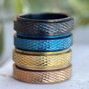 Peut inclure: Une pile de cinq bagues métalliques de couleurs variées : noir, bleu, or et or rose. Chaque bague présente une bande texturée à motif de losanges. Les bagues sont empilées les unes sur les autres, mettant en valeur leurs différentes teintes et leurs motifs.