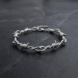 Może przedstawiać: Srebrna bransoletka z motywem drutu kolczastego. Metalowa bransoletka posiada ostre, spiczaste kolce. Bransoletka jest prezentowana na ciemnej, teksturowanej powierzchni.