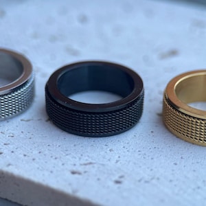 Puede incluir: Tres anillos en fila, cada uno con un acabado diferente. El anillo central es negro con una textura de malla, flanqueado por un anillo plateado a la izquierda y un anillo dorado a la derecha. Los anillos están sobre una superficie clara y moteada.