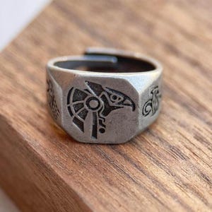 Puede incluir: Un anillo plateado con un diseño de pájaro grabado y patrones geométricos. El anillo ajustable se encuentra sobre una superficie de madera, mostrando su detallada artesanía y estética vintage. El diseño del anillo presenta un ave rapaz.