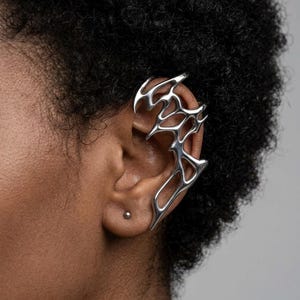 May include: A silver ear cuff with an intricate, openwork design. The ear cuff wraps around the ear, featuring a series of interconnected, abstract shapes. The jewelry is made of a shiny, metallic material.